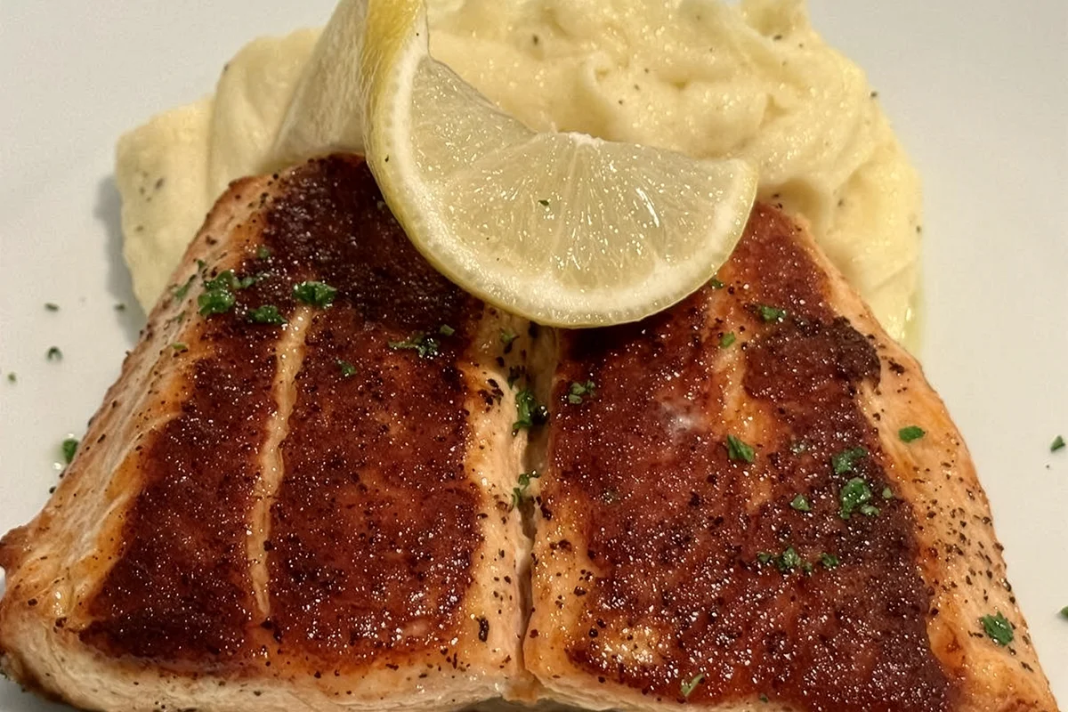 Two pieces of pan-seared salmon with a crispy golden crust, served with creamy mashed potatoes and a lemon garnish.