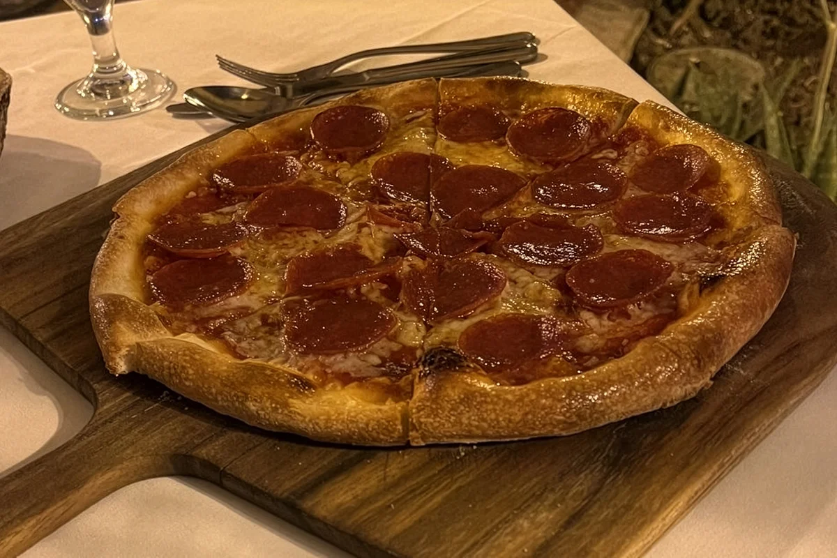 A whole pepperoni pizza with a crispy, charred crust served on a wooden cutting board in a dimly lit restaurant setting.
